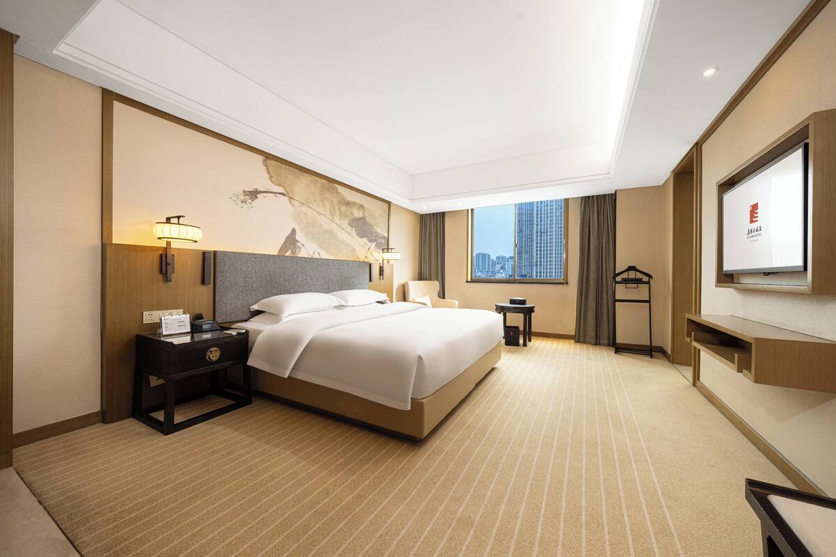 Ein Hotelzimmer mit einem großen Bett, einem Nachttisch, einem Schreibtisch, einem Sessel und Blick auf die Stadt.