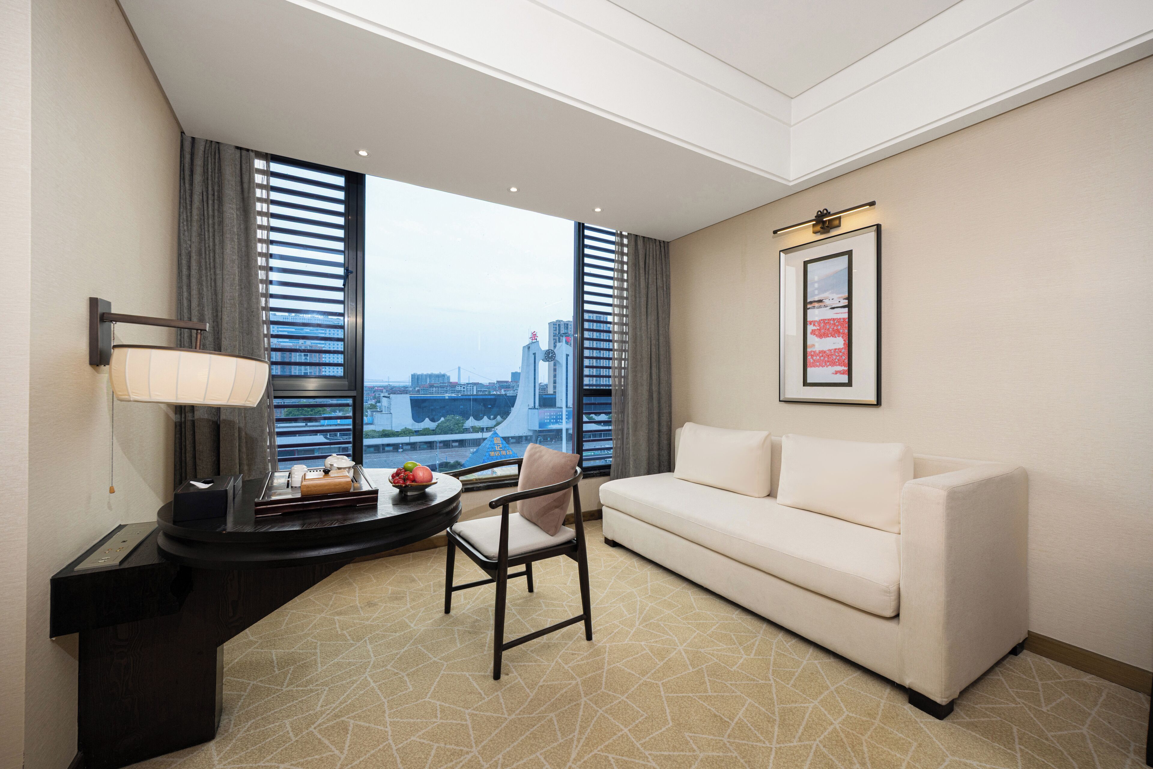 A hotel room with a white sofa, a desk with a lamp, a chair, and a large window with blinds.