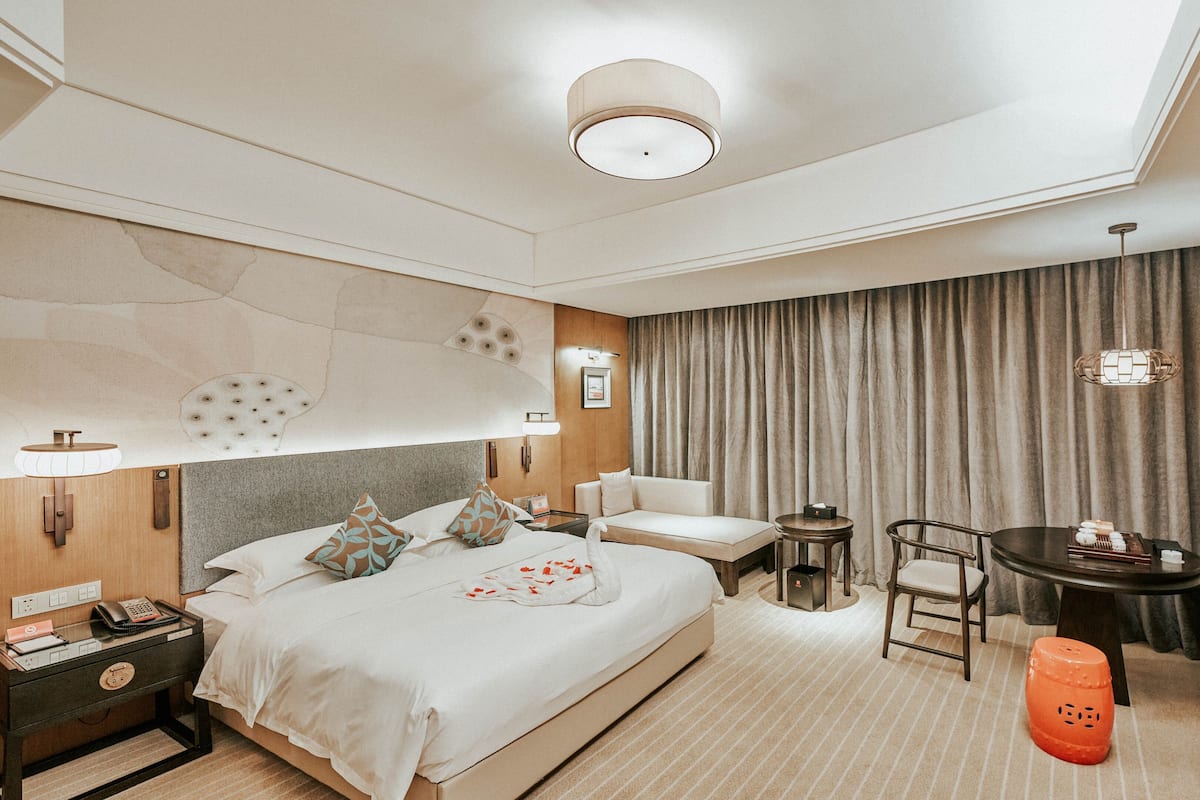 Ein Hotelzimmer mit Bett, Nachttisch, Lampe, Sessel, Fernseher und kleinem Tisch mit Lampe.