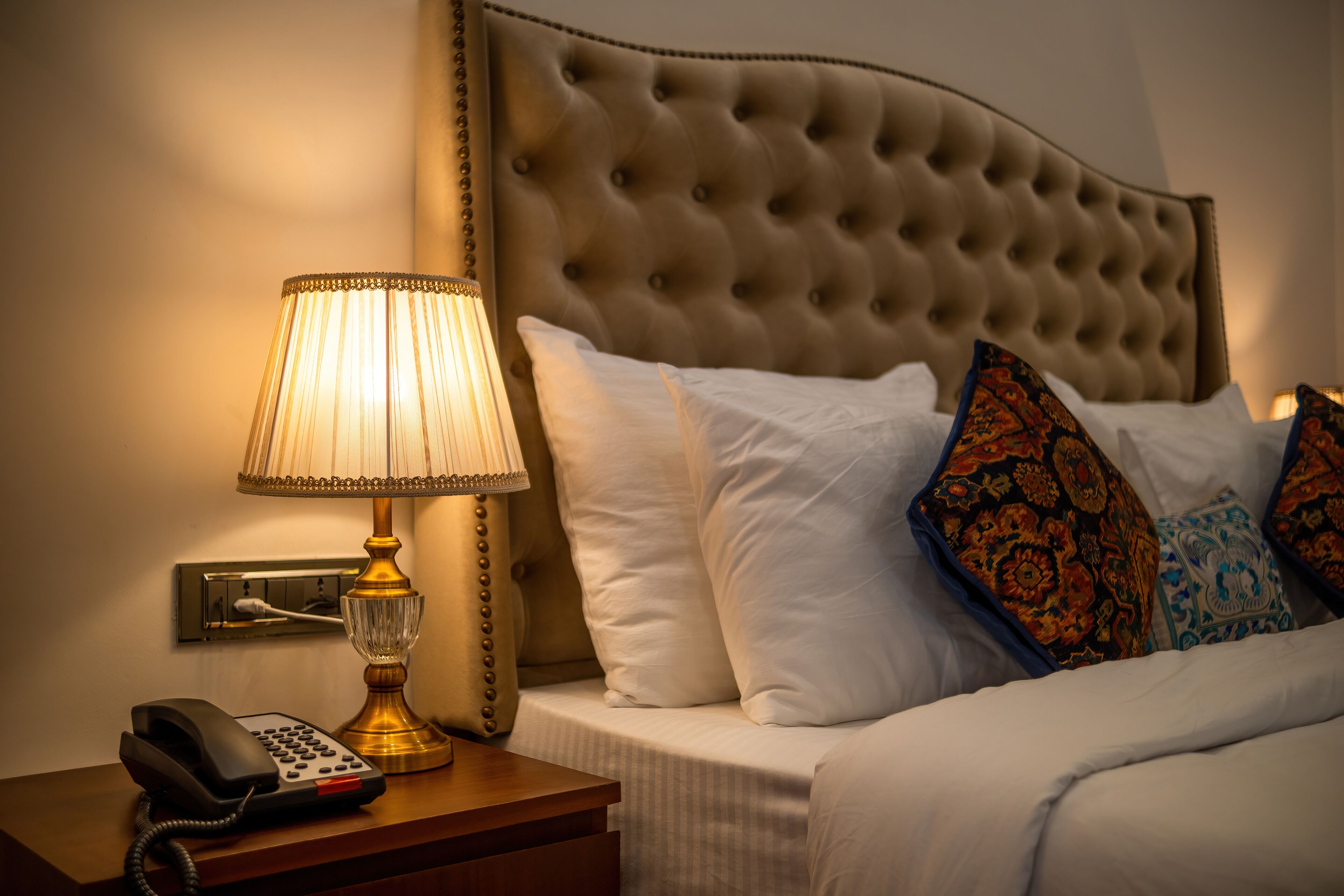 A hotel room with a tufted headboard, a bedside lamp, a telephone, and a bed with white and patterned pillows.