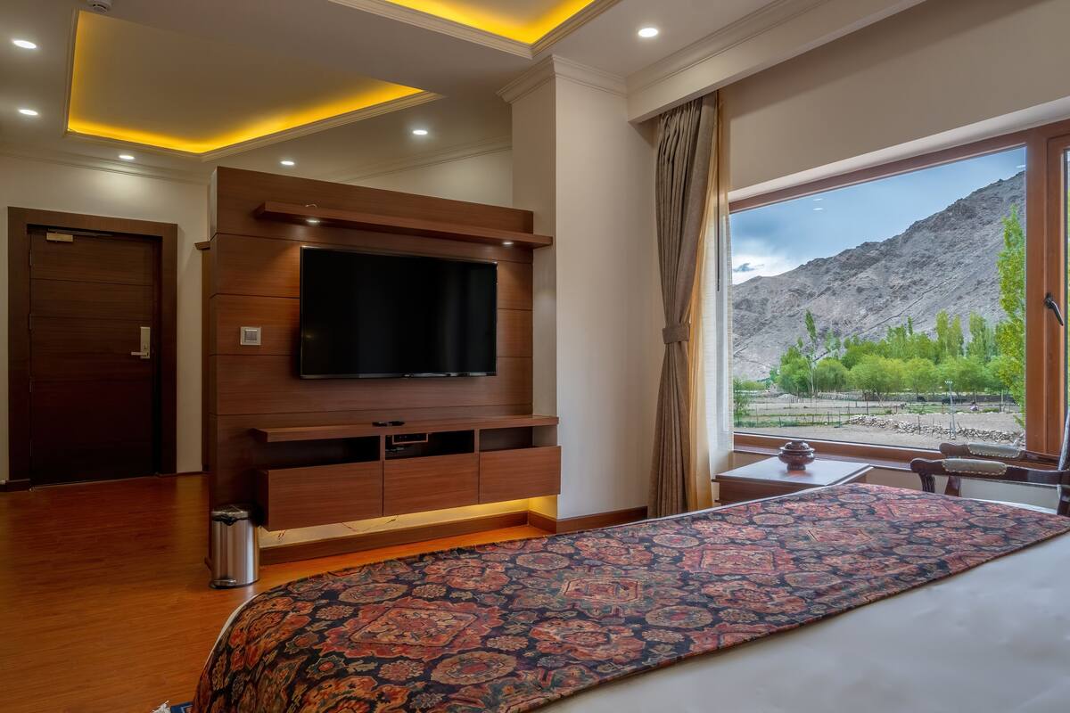 A hotel room with a large bed, a flat-screen TV mounted on a wooden cabinet, a small table with a vase, and a view of mountains through a large window.