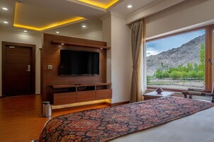 Habitación de hotel con una cama grande, un televisor de pantalla plana montado en un mueble de madera, una mesita con un florero y vistas a montañas a través de un ventanal amplio.
