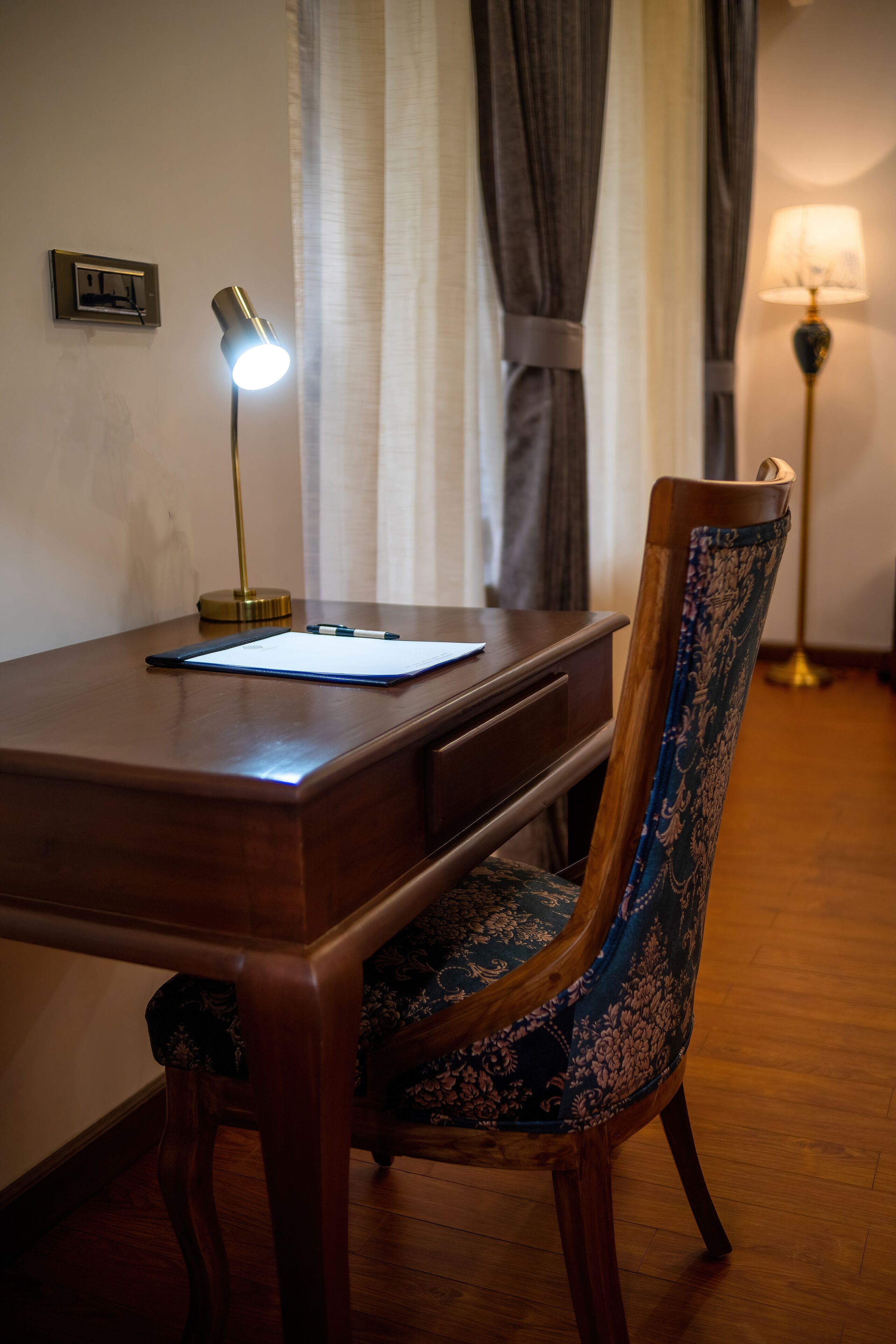 Un escritorio de madera con una lámpara, una silla con un cojín estampado y un enchufe de pared.