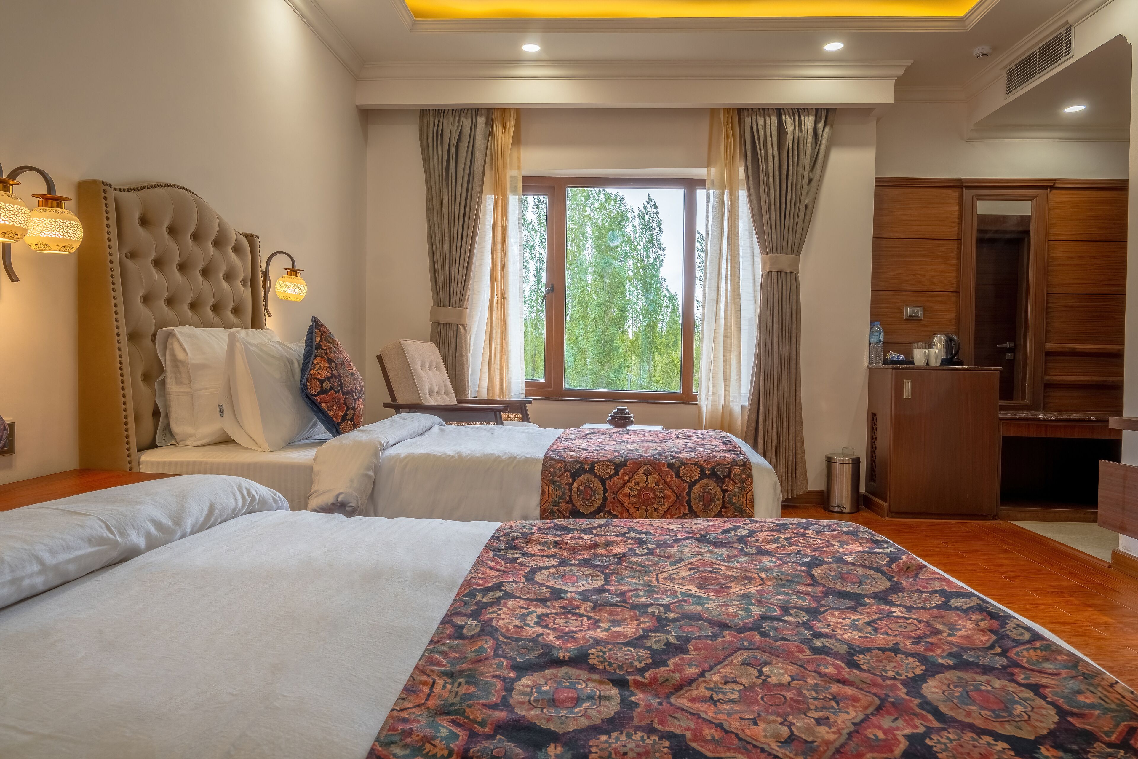 A hotel room with two beds, a large window with curtains, a wardrobe, and a small table with a kettle.