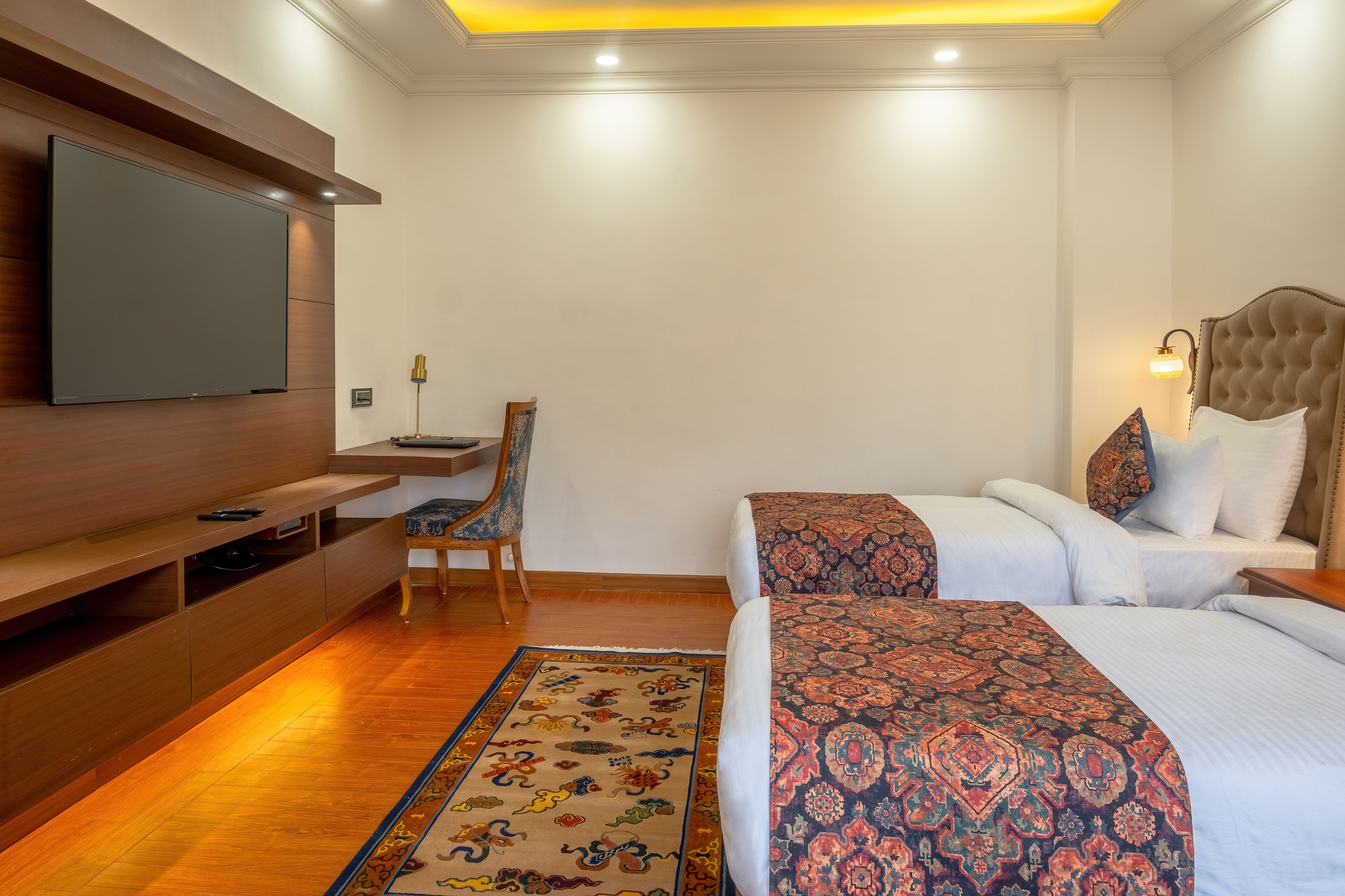 A hotel room with a flat-screen TV, a desk with a chair, two beds with patterned bedspreads, and a decorative rug.