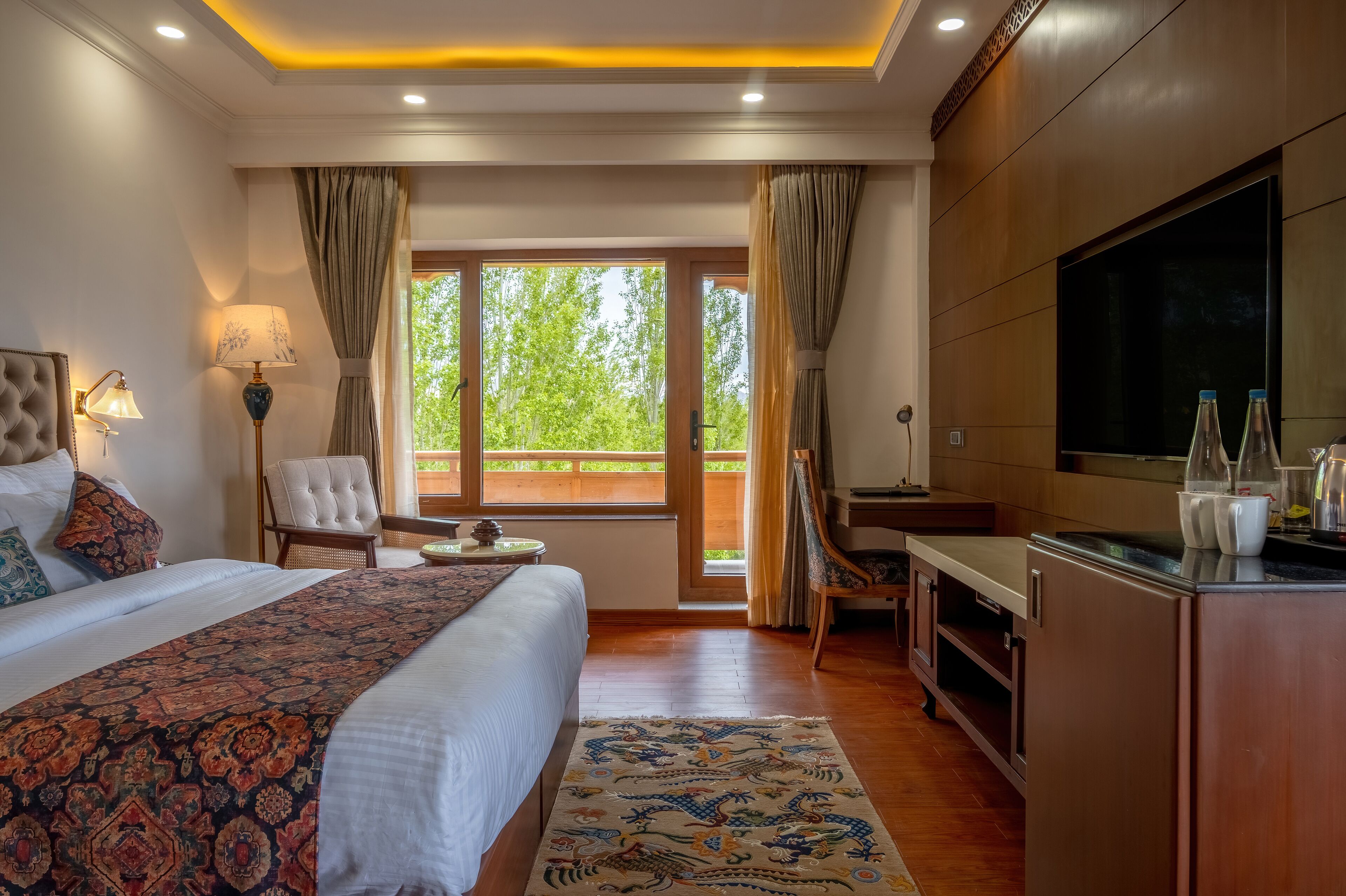 A hotel room with a large bed, a desk, a television, and a view of greenery outside.