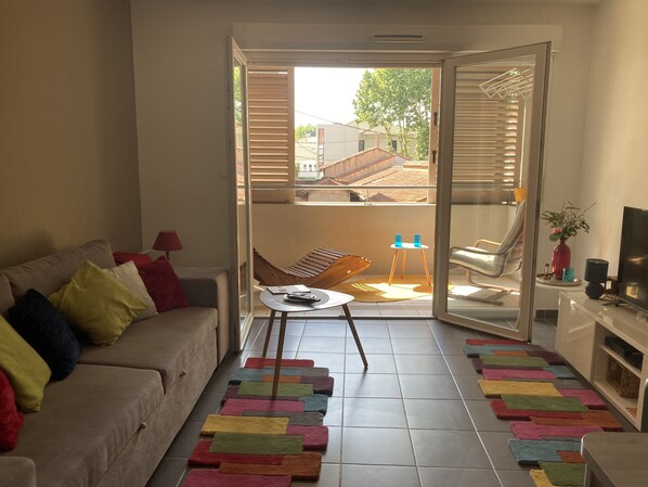 A living room with a sofa, a coffee table, and a TV on a stand. There is a balcony with a chair and a table.