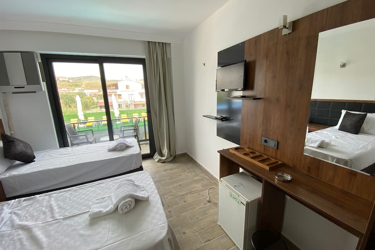 A hotel room with two beds, a wooden desk, a small refrigerator, a TV mounted on the wall, and a balcony with a view of buildings and greenery.
