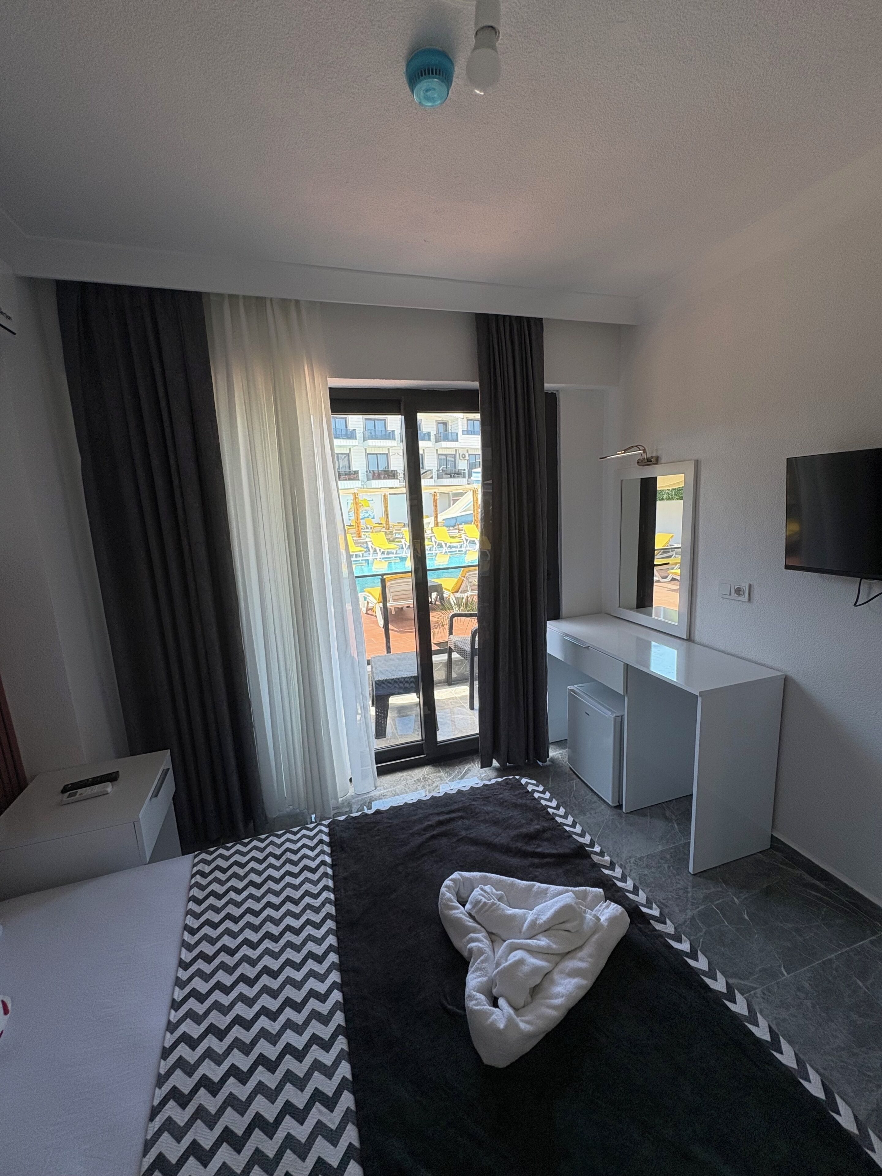 Ein Hotelzimmer mit Bett, Schreibtisch, Fernseher und Blick auf einen Außenbereich mit Stühlen und Tischen.