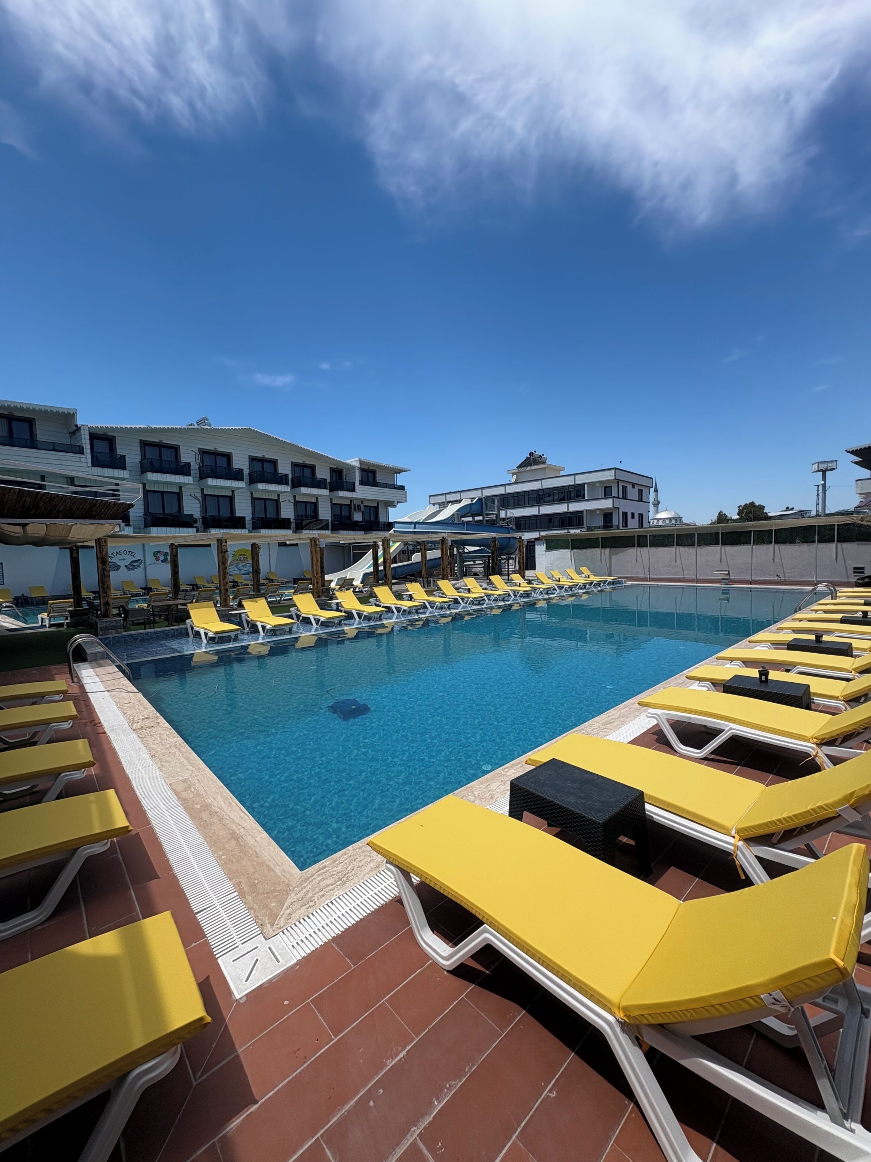 Una piscina con sillas de descanso amarillas, una terraza con baldosas y edificios al fondo.