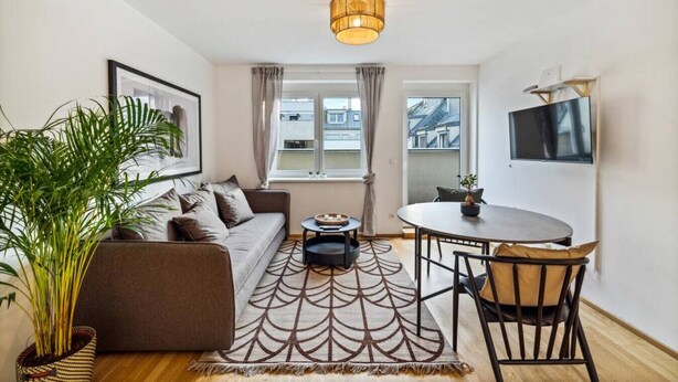 A modern living room with a sofa, a coffee table, a dining table with chairs, a large potted plant, and a television mounted on the wall. - Botanical Design Apart in Erlachplatz (Vienna)