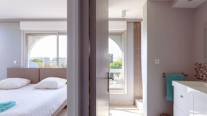 Une chambre d’hôtel moderne, dotée d’un lit, d’une salle de bain avec un meuble-lavabo blanc et offrant une vue sur la ville grâce à de grandes fenêtres.
