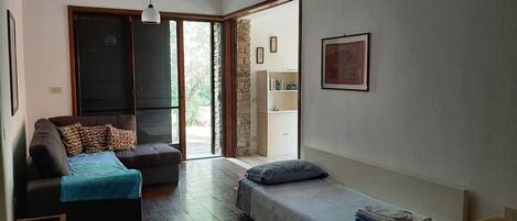 A compact room with a sofa, a small bed, a wooden stool, and a door leading to a balcony.