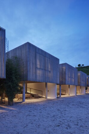 A modern building with a series of rectangular structures, possibly a hotel or resort, with a sandy ground in front and a clear sky.