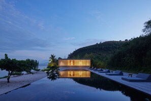 A modern building with a glass facade, a wooden deck, and lounge chairs by a calm body of water.