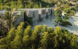 Um edifício moderno com design arquitetônico único, rodeado por vegetação exuberante e uma área arenosa.