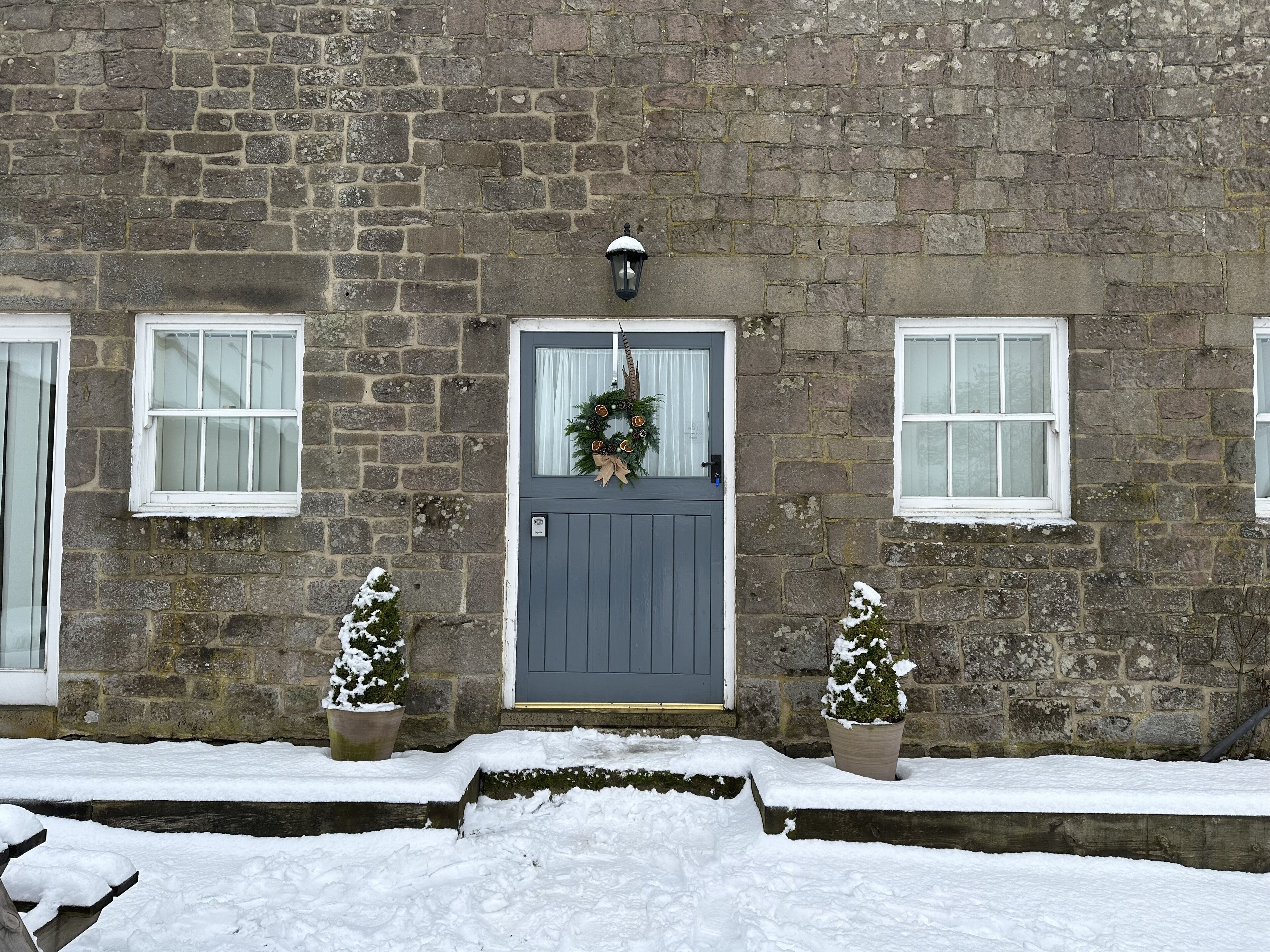 A stone building with a grey door, two windows, and two potted plants with snow on them.