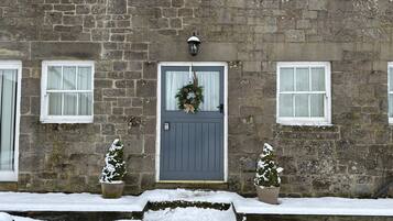 A stone building with a grey door, two windows, and two potted plants with snow on them.