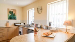 Una cocina con encimera de granito, una botella de vino, vasos y un plato de comida. Se observa una lavadora, una cafetera y un reloj en la pared.