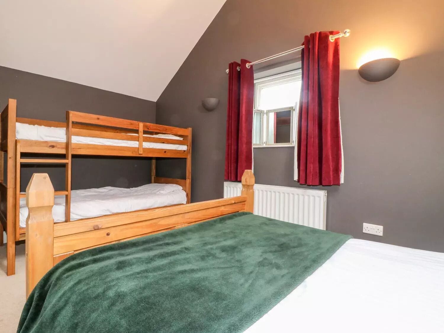 A room with a bunk bed, a single bed, a window with red curtains, a wall-mounted light, and a radiator.