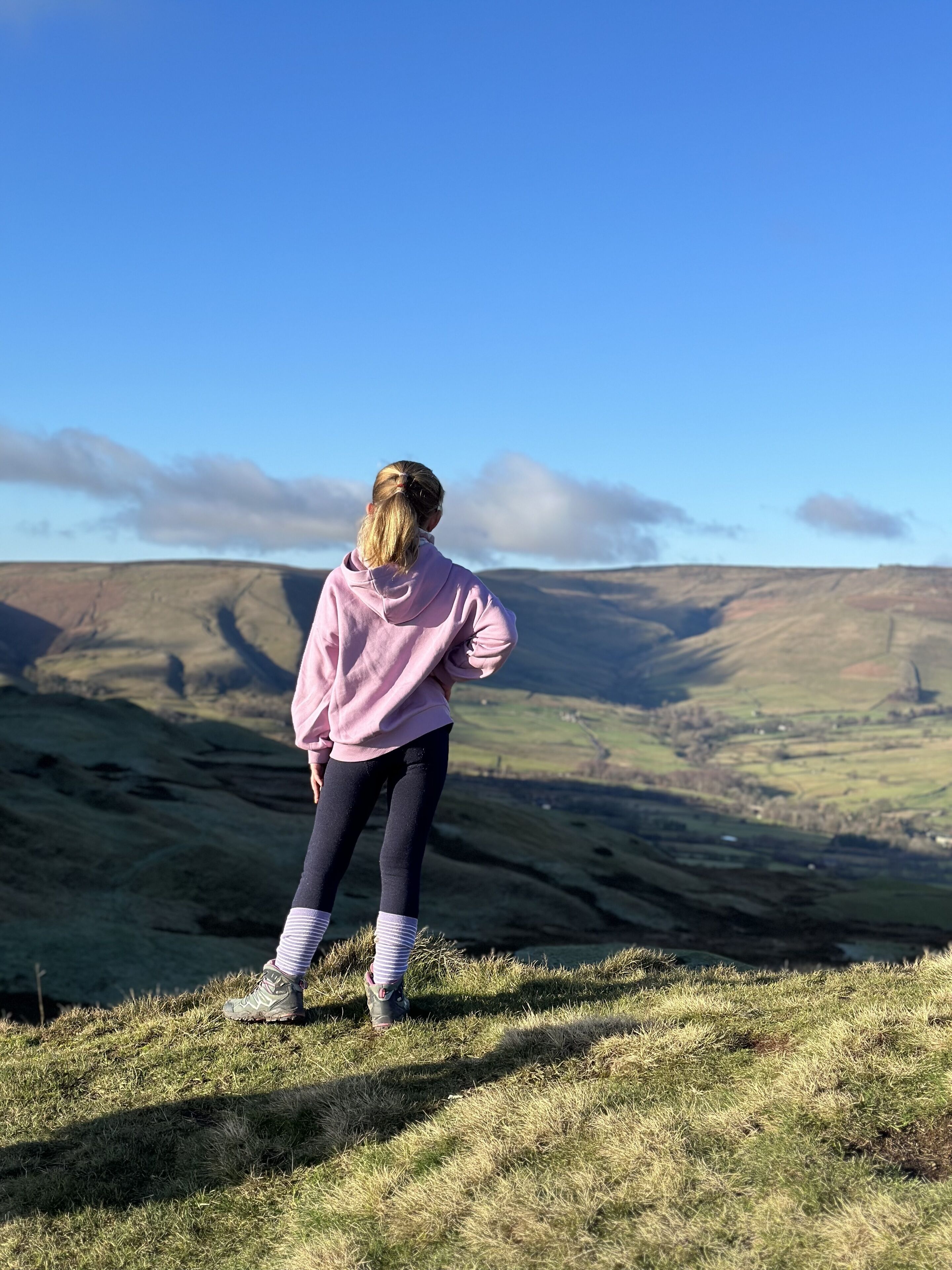 A person standing on a grassy hill overlooking a valley with rolling hills and a clear blue sky.