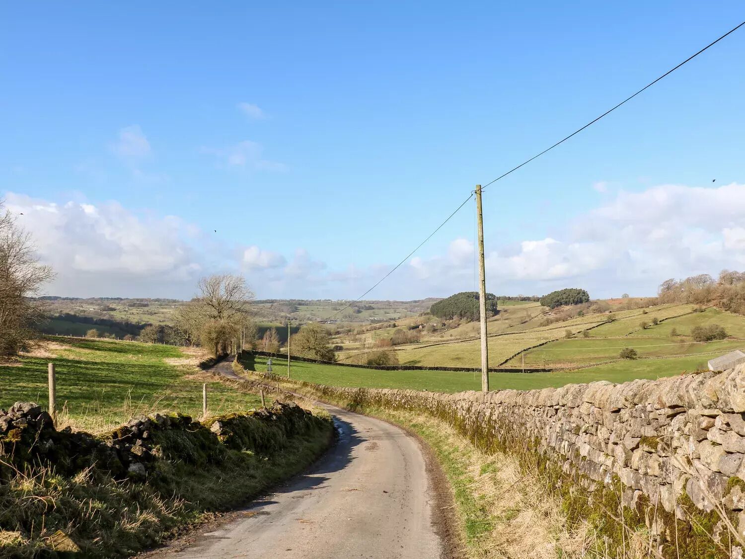 A rural road flanked by stone walls and greenery, leading through a hilly landscape.