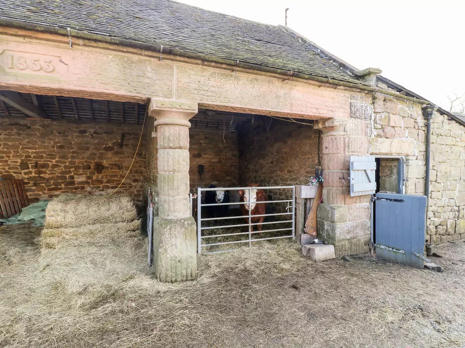 A stone building with a date '1853' carved into the wall, a metal gate, and hay bales outside.