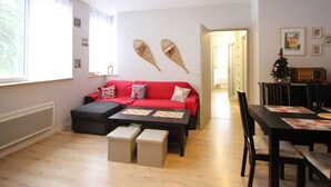 Una sala de estar con un sofá rojo, una mesa de centro y una mesa de comedor con sillas. Hay una ventana con cortinas, una puerta que da a otra habitación y un pequeño árbol de Navidad sobre la mesa de comedor.