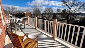 A balcony with a wooden bench, white railing, and a view of a building and trees. - Scenic South Main Apartment with Breathtaking Views for 3 Guests (Saint Charles)