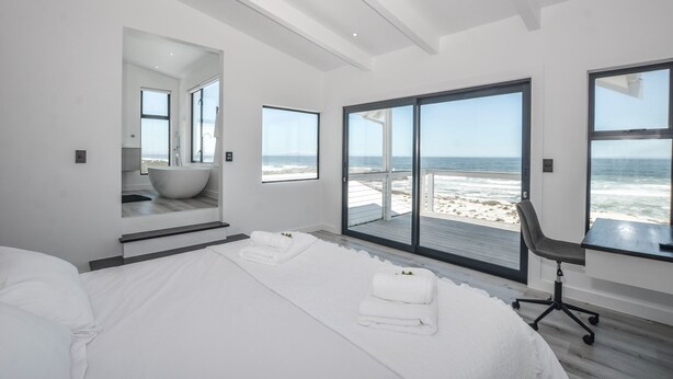 A modern bedroom with a large bed, a bathroom with a bathtub, and a view of the beach. - Sandbanks Villa (Yzerfontein)