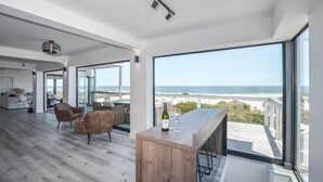 A spacious living area with a wooden floor, a bar area with a bottle and glasses, and a large window overlooking the ocean. - Sandbanks Villa (Yzerfontein)