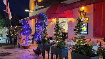 A hotel exterior with a red awning, outdoor seating area, and Christmas trees with lights.