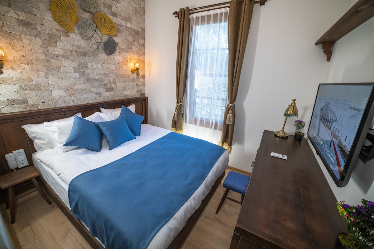 A hotel room with a brick wall, a bed with blue bedding, a wooden nightstand, a desk, and a wall-mounted TV displaying a Poland map.