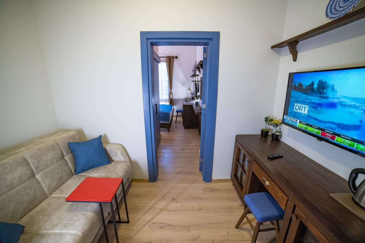 A living room with a sofa, a red table, a wooden desk, and a TV displaying a weather forecast.