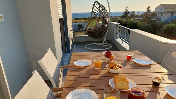 A wooden table set for breakfast with plates, glasses of orange juice, a bowl of cookies, and a jar of jam on a balcony overlooking the sea. - Elia Luxury Apartments Estd 2022 (Preveza)