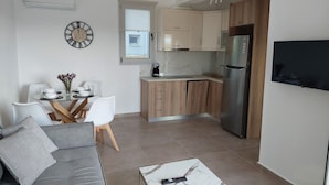 A modern living room with a grey sofa, a coffee table, and a dining area with a white table and chairs. A kitchen with wooden cabinets and a stainless steel refrigerator is visible in the background. - Elia Luxury Apartments Estd 2022 (Preveza)