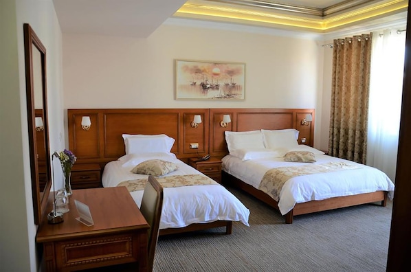 A hotel room with two beds, a desk, and a chair. There is a painting on the wall and a window with curtains.