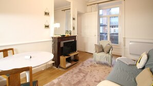 A living room with a fireplace, a dining table, a sofa, and a window with curtains. - Mont-Dore Center: Beautiful T2 Apartment with Wifi Near the Thermal Baths (Mont-Dore)