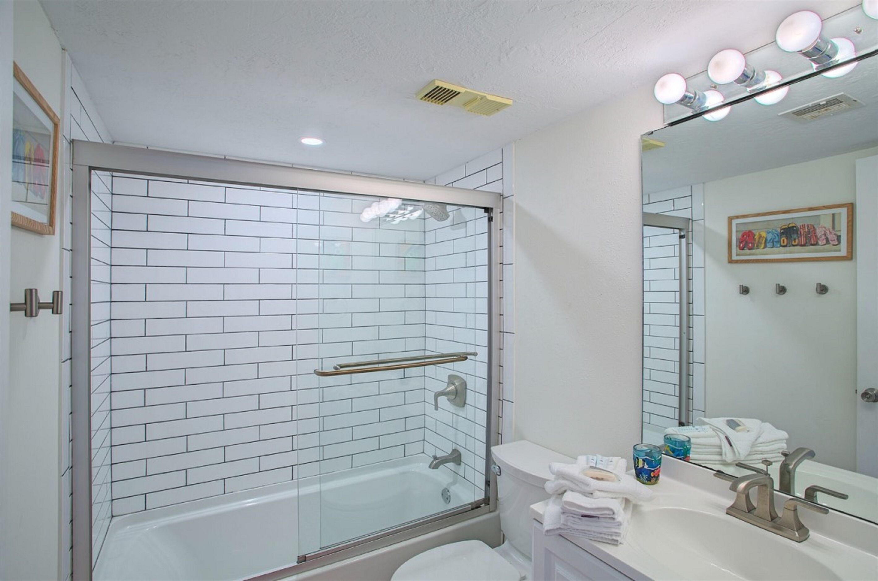 A modern bathroom with a glass-enclosed shower, white tiled walls, a white sink, and a mirror with lights above it.