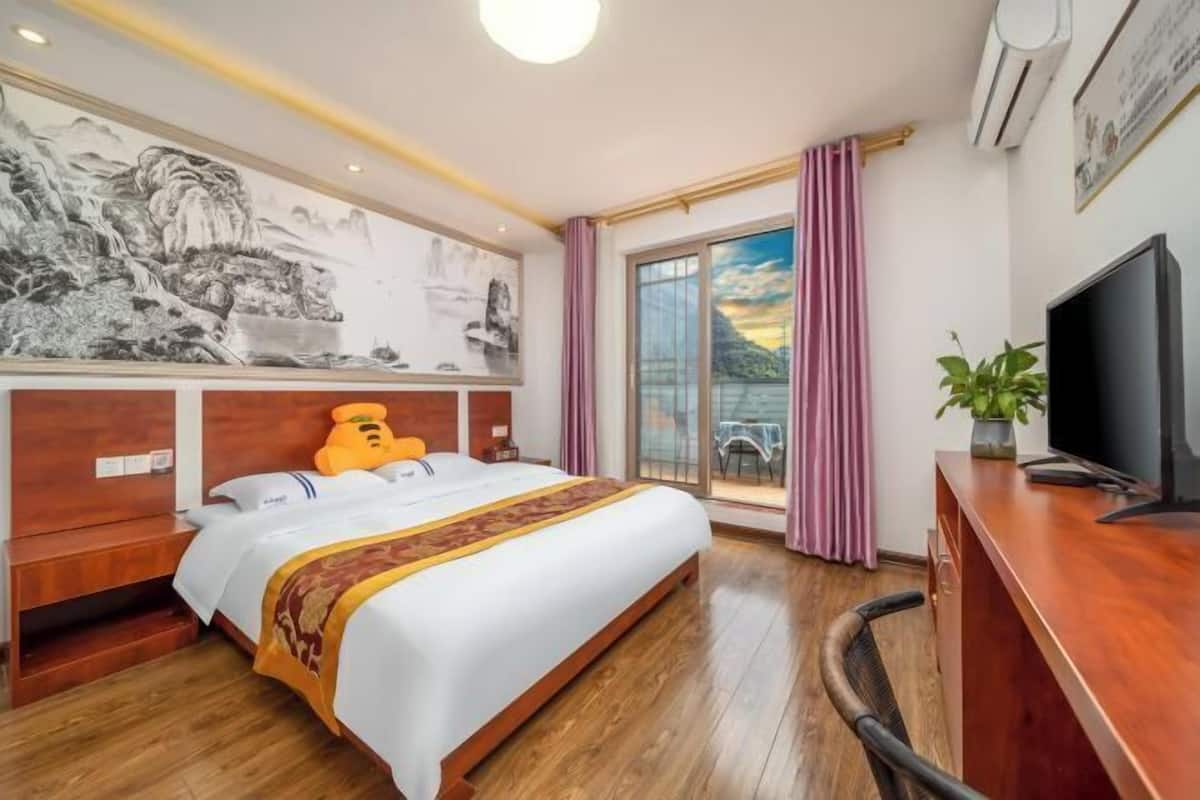 Habitación de hotel con una cama grande, cabecera de madera, televisor, escritorio y vistas a un balcón con cortinas.