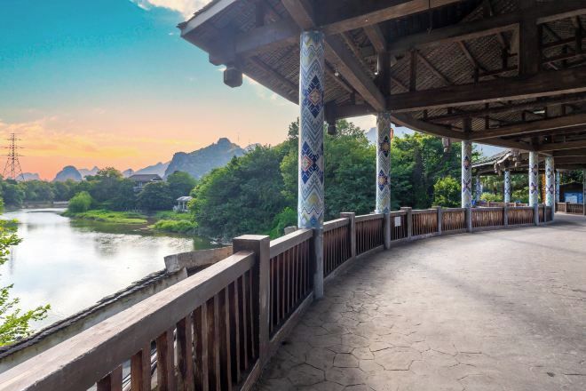 A covered walkway with patterned columns and a wooden railing, overlooking a calm river and lush greenery.
