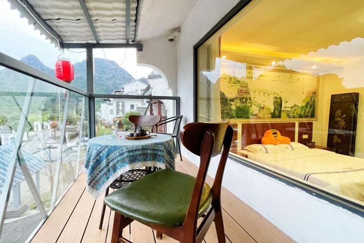 A balcony with a table and chairs, a bed with a view of mountains, and a large window with artwork.