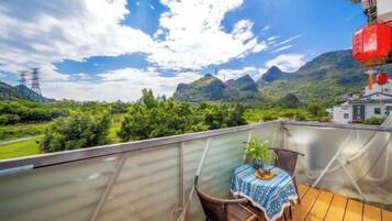 A balcony with a table and chairs, overlooking a mountainous landscape with greenery and a building in the distance.