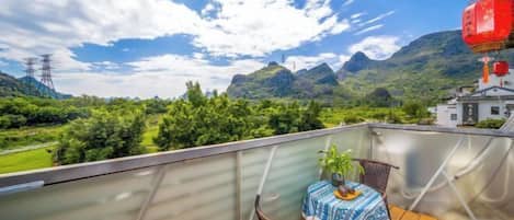 A balcony with a table and chairs, overlooking a mountainous landscape with greenery and a building in the distance.