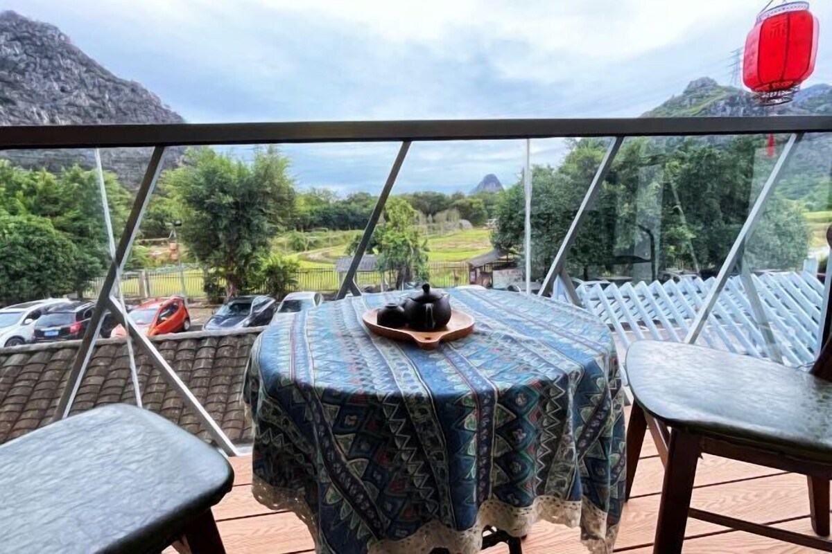 A balcony with a table covered in a patterned tablecloth, a teapot, and a tray. The balcony overlooks a mountainous landscape with greenery and parked cars.