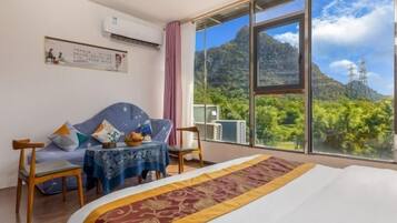 A hotel room with a bed, a sofa, a small table, and a view of mountains and greenery.