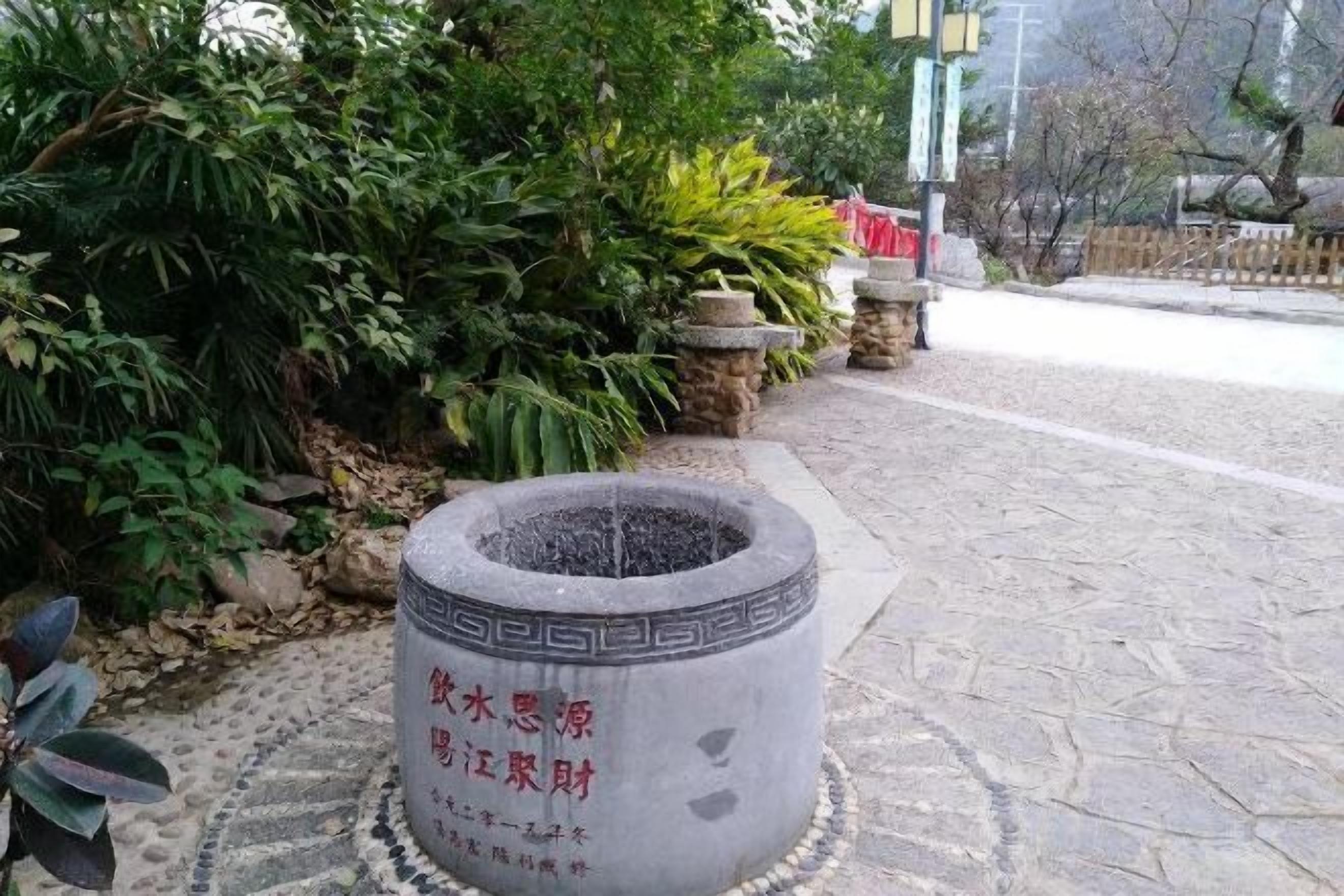 Un bac à fleurs en pierre orné d’inscriptions, entouré de verdure et bordé d’un chemin pavé.