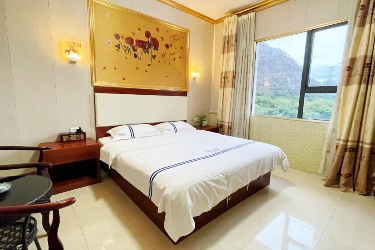 A neatly arranged hotel room with a large bed, a bedside table, a chair, and a window offering a view of a mountainous landscape.