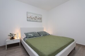 Een slaapkamer met een groen geborduurde spreidekking, een witte nachtkastje met een lamp en een bloemig kunstwerk aan de muur.