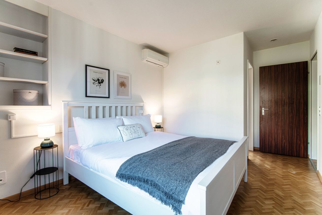 Ein Schlafzimmer mit einem weißen Bett, Nachttischen mit Lampen, einer Wandklimaanlage und einem hölzernen Kleiderschrank.